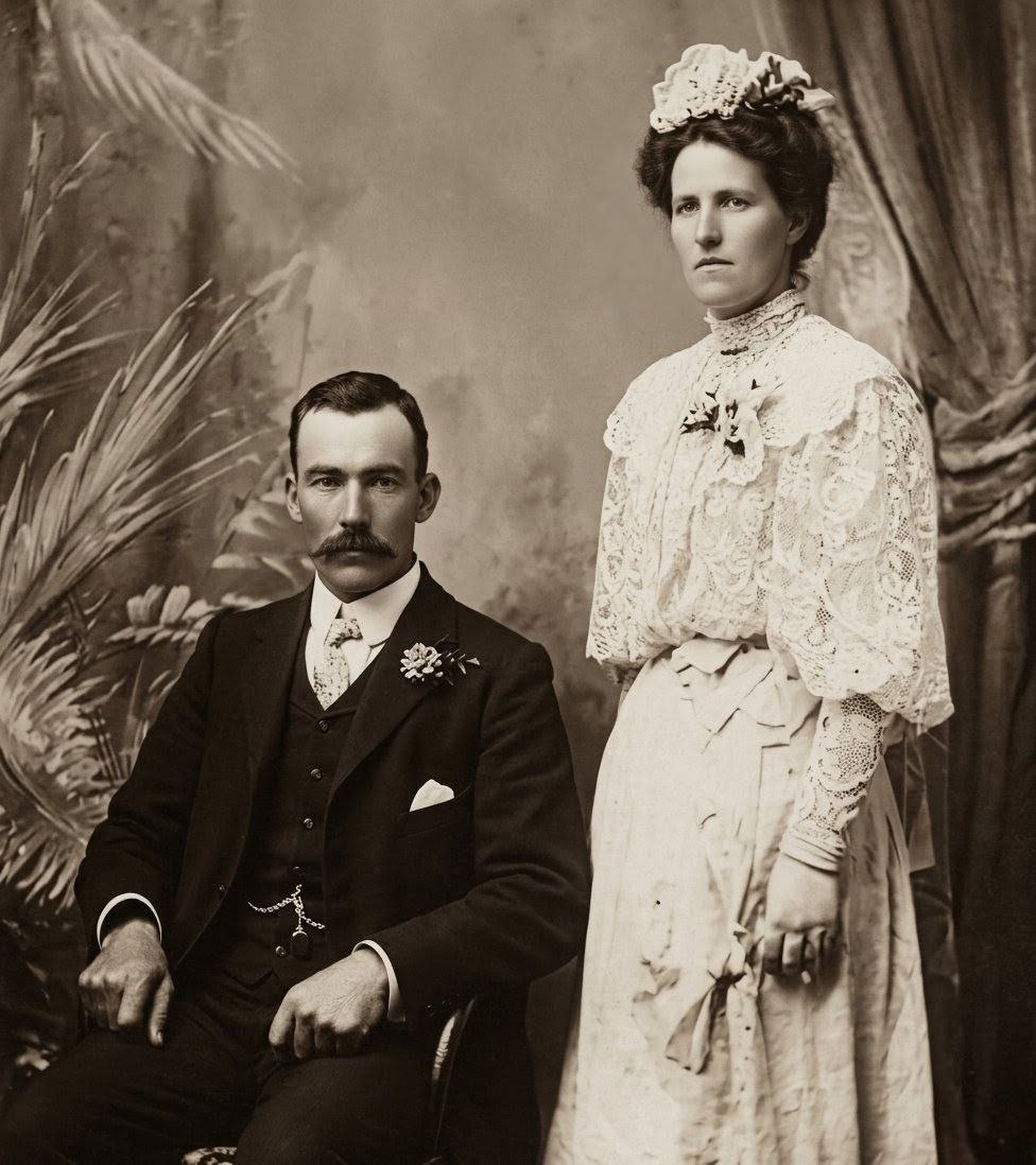 Francis Charles 'Charlie' Telfer and Amy Ellen Provis on their wedding day, 6 April 1909. Studio portrait, sepia tones. Charlie (aged 33) in dark three-piece suit with handlebar mustache, white pocket square, watch chain, and boutonnière. Amy (aged 25) in elaborate white lace gown with high ruffled collar, voluminous sleeves, and floral headpiece, holding a small bouquet. Painted tropical foliage backdrop and draped curtain.