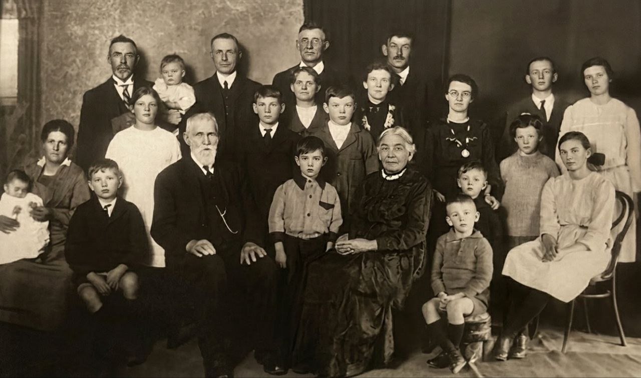 Telfer family reunion, circa 1920. Large multi-generational group portrait of ~25 individuals. John Telfer (1847–1929, aged ~73) and Caroline Amelia Telfer (1842–1929, aged ~78) seated center with white beard and lace collar respectively. Their son Francis Charles 'Charlie' Telfer (1875–1954, aged ~45) and wife Amy Ellen Telfer (1884–1951, aged ~36) among the standing adults with their children and grandchildren. Studio setting with textured wallpaper backdrop.